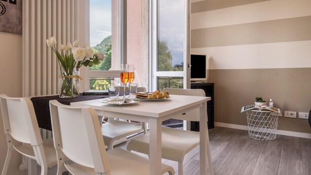 Ein modernes Zimmer mit Esstisch, weißen Stühlen, einer Vase mit Blumen, einem kleinen Beistelltisch mit Pflanze und einem großen Fenster mit Blick ins Grüne.