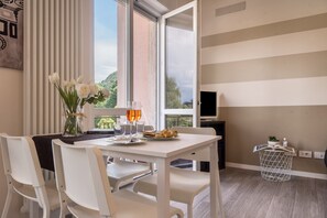 Une pièce moderne comprenant une table à manger, des chaises blanches, un vase avec des fleurs, une petite table d’appoint avec une plante, et une grande fenêtre donnant sur un paysage verdoyant.