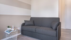 Ein graues Sofa mit zwei Kissen, ein weißer Nachttisch mit einer Pflanze und einer Zeitschrift, sowie eine Wand mit horizontalen Streifen.