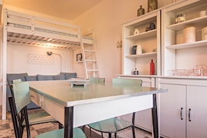 A compact room with a bunk bed, a dining table with chairs, a kitchenette, and a shelving unit with various items.