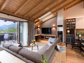 A modern living room with a large sofa, a dining area, and a kitchen. There is a balcony with a view of mountains.