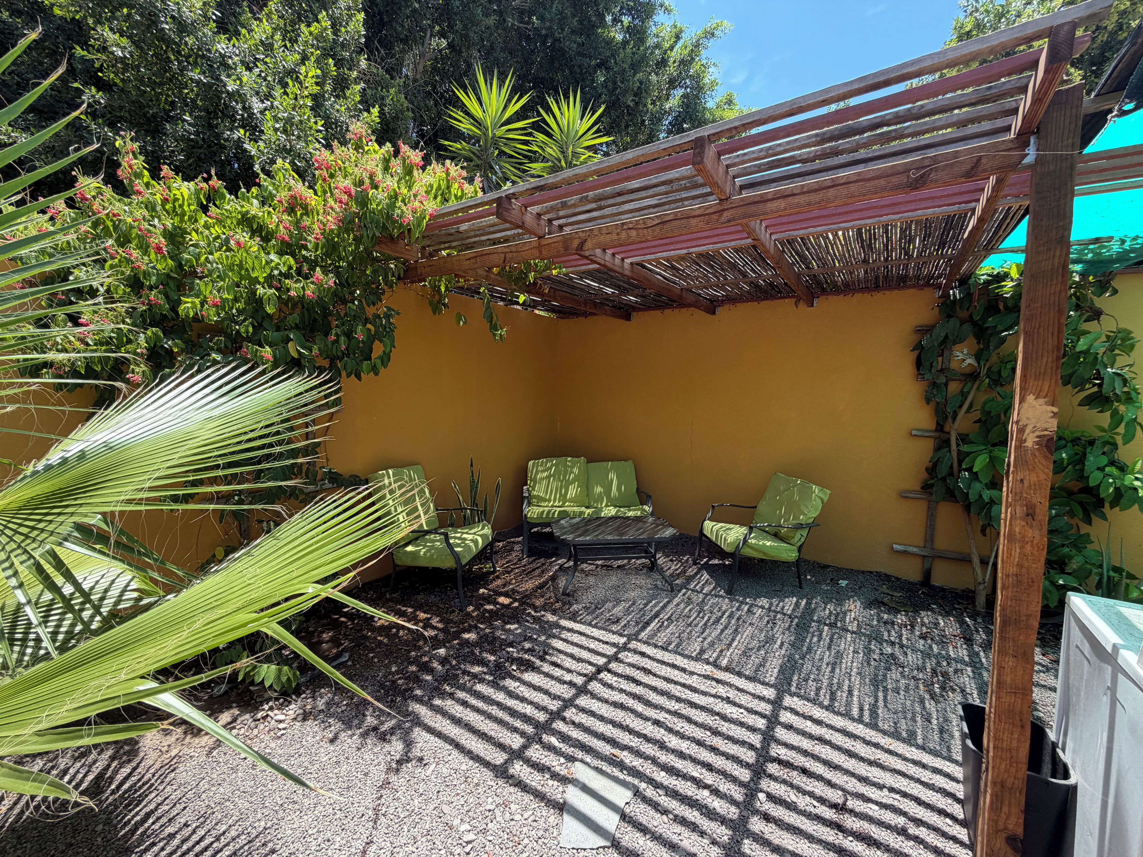 Un espace extérieur aménagé avec une zone de repos couverte, comprenant deux chaises et une table, le tout entouré de verdure et adossé à un mur jaune.