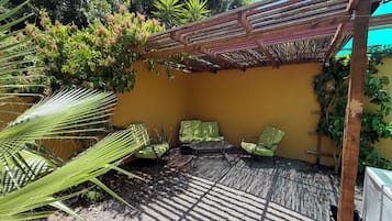 Un espace extérieur aménagé avec une zone de repos couverte, comprenant deux chaises et une table, le tout entouré de verdure et adossé à un mur jaune.