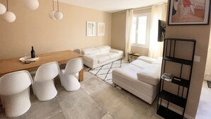 A modern, well-lit room with a wooden dining table, white chairs, a white sofa, and a black metal chair. There is a framed picture on the wall and a window with curtains. - Villa Alexandra (Le Crotoy)