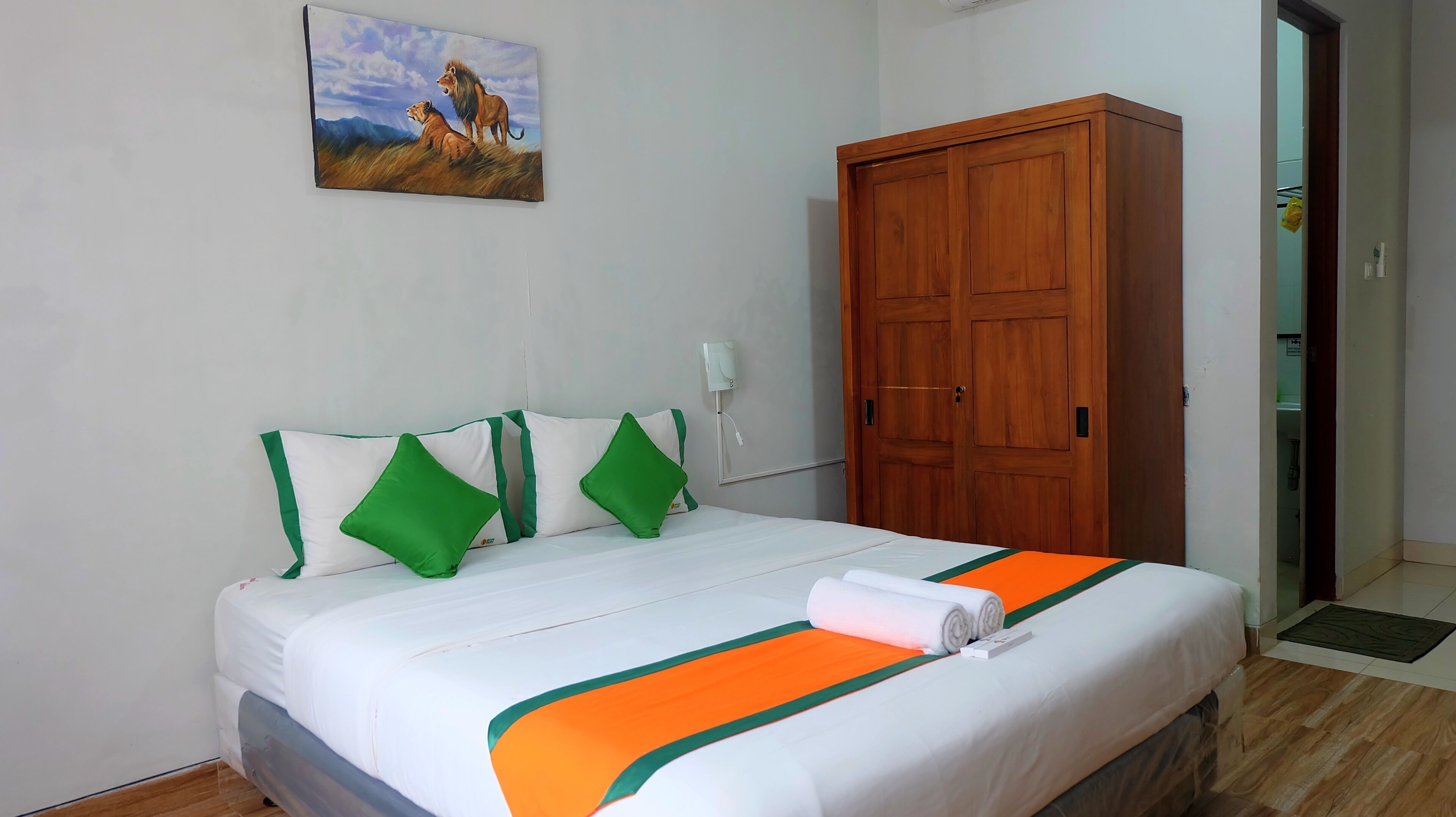 A hotel room with a bed, green and white pillows, a wooden wardrobe, and a painting of lions on the wall.