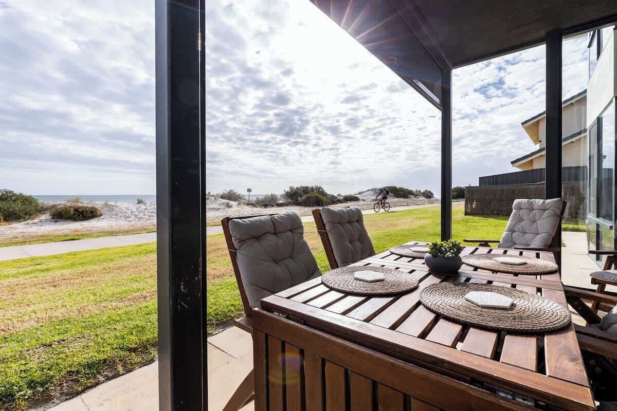 A wooden table with four chairs, a potted plant, and woven placemats on a patio with a view of the beach and ocean.