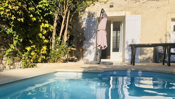 A swimming pool with a stone edge, surrounded by greenery and a building with a door and windows. - LA Villa Avignon - Centre Ville - Piscine Privée - Intramuros (Avignon)