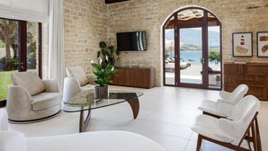 A spacious living room with a stone wall, a glass coffee table, and a view of the sea through large glass doors.