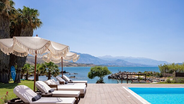 A poolside area with lounge chairs and umbrellas, overlooking a body of water and mountains.