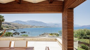 A wooden deck with a table and chairs, overlooking a coastal landscape with mountains and a body of water.