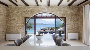A dining room with a long table, stone walls, and a view of the sea.