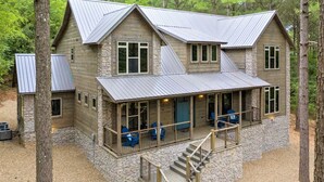 A two-story log cabin with a stone foundation, a covered porch with outdoor seating, and a staircase leading to the upper level. - Forest Home|5B/5.5BA|Hot tub|Game Room|Playset (Broken Bow)