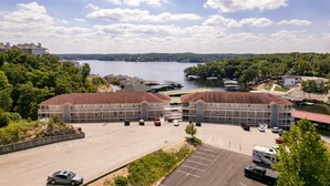 Ein großer Parkplatz mit mehreren Autos und Wohnmobilen, ein mehrstöckiges Gebäude mit rotem Dach und ein See mit Booten im Hintergrund.