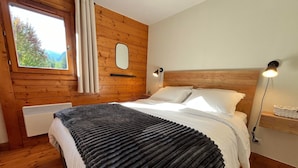 A wooden-paneled bedroom with a bed, a window, a mirror, and a bedside table with a lamp. - T3 Apartment in Samoëns Center with Balcony and Private Parking (Samoëns)