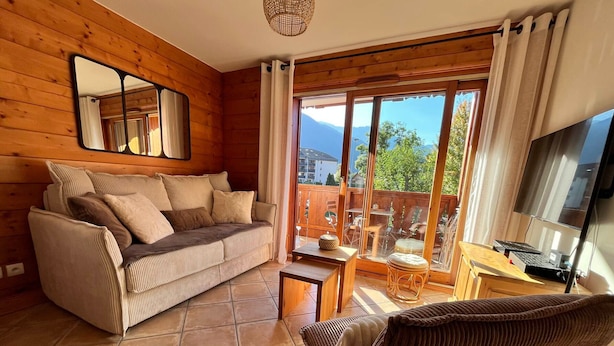 A cozy room with a wooden interior, a sofa, a small table, a chair, and a TV. - T3 Apartment in Samoëns Center with Balcony and Private Parking (Samoëns)
