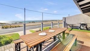 A wooden deck with tables and chairs overlooking a coastal road and ocean. - Queensland South - Aldinga Beach - C21 SouthCoast Holidays (Aldinga Beach)
