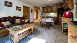 A cozy living room with a wooden sofa, a coffee table, and a dining area with a table and chairs. The kitchen has wooden cabinets and a red microwave.