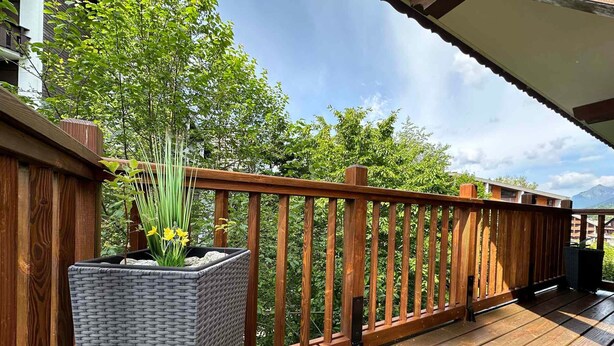 A wooden deck with a railing, a planter box with plants, and a view of trees and mountains in the background.