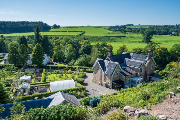 A stone house with a sloped roof and solar panels, surrounded by lush green fields and trees.
