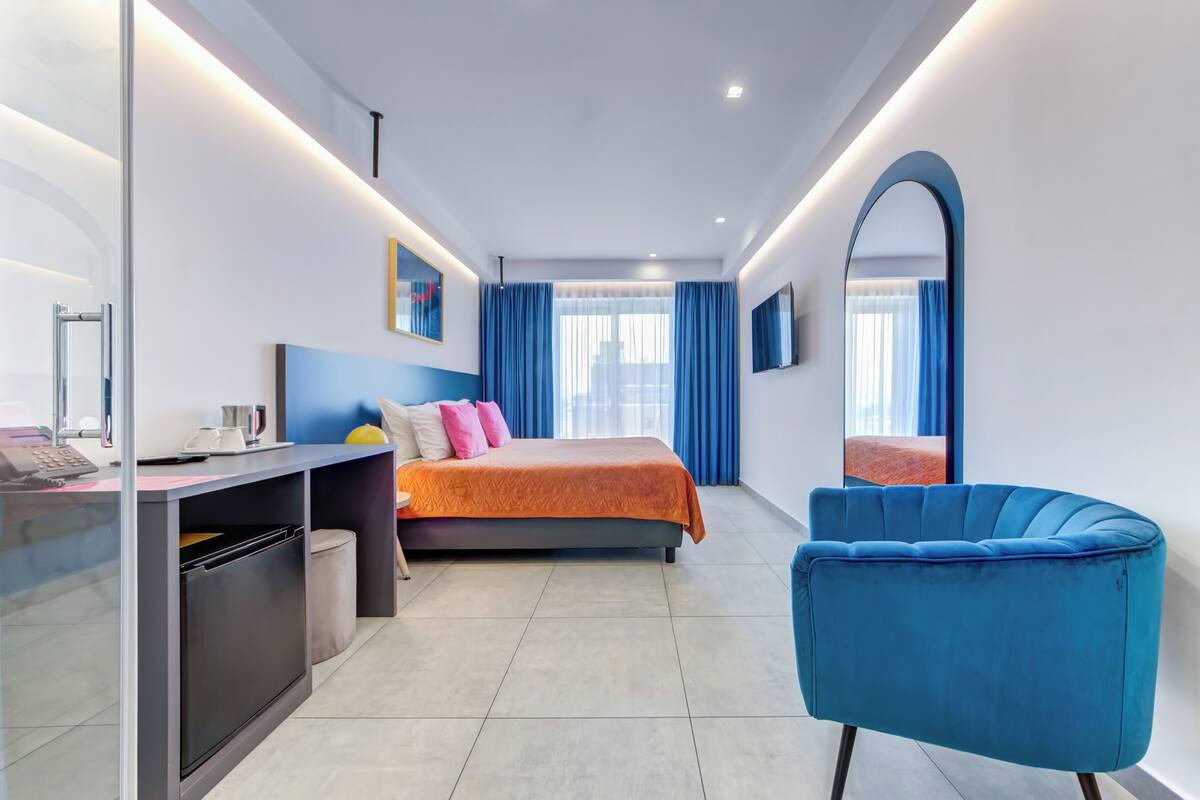 A modern hotel room with a bed, a desk with a computer, a chair, and a large mirror.