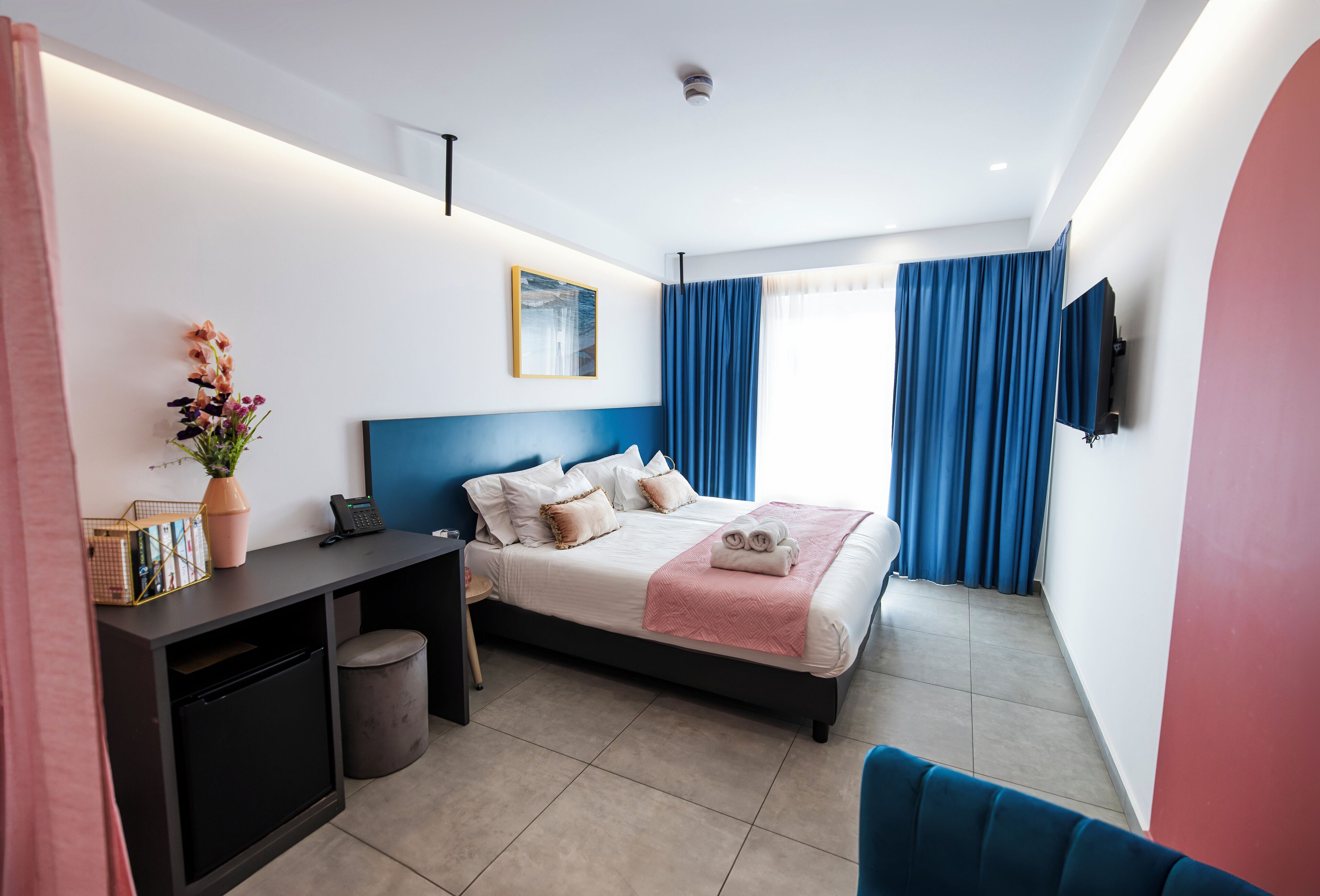 A modern hotel room with a bed, desk, and chair, a small trash bin, and a TV mounted on the wall.