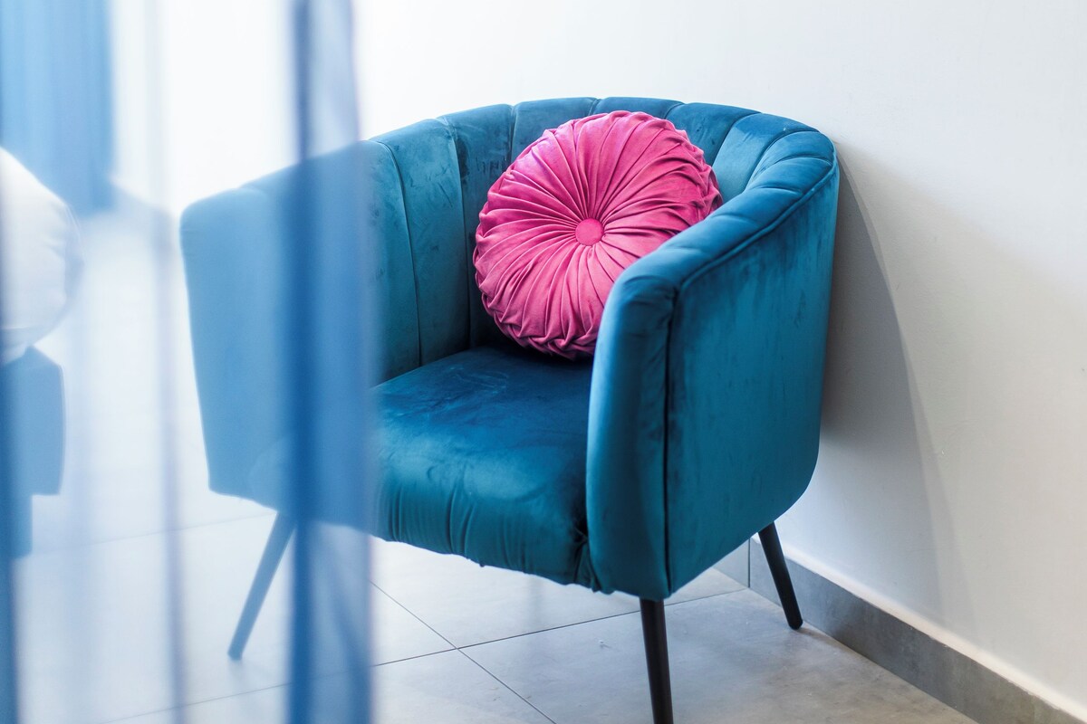 A corner room with a blue armchair, a pink pillow, and a wall-mounted TV.