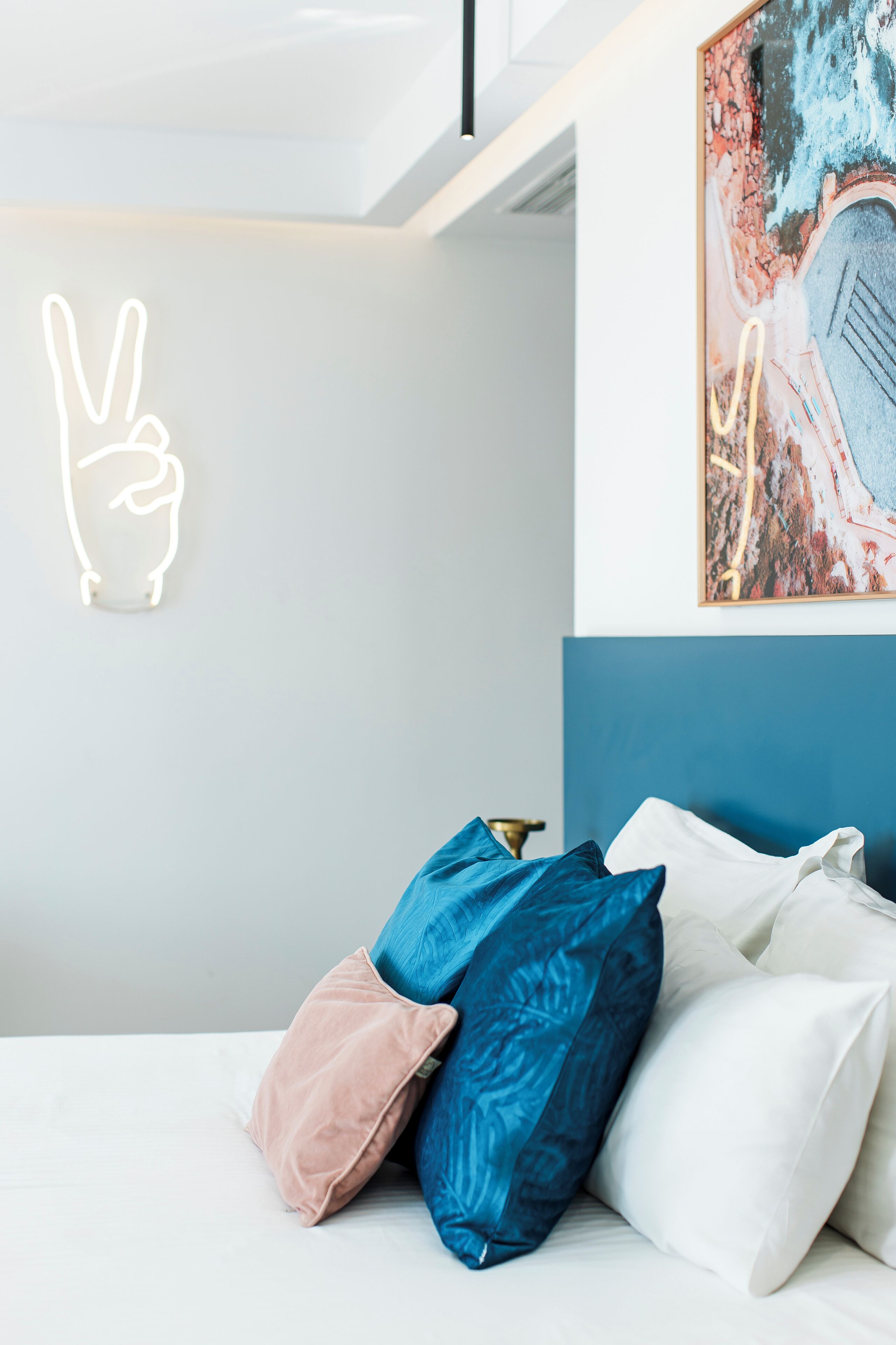 A modern bedroom with a bed, pillows, a neon peace sign, and a colorful abstract painting on the wall.