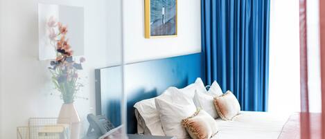 A hotel room with a bed, a bedside table, a phone, a vase with flowers, a framed picture on the wall, and a blue curtain.