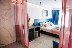 Ein modernes Hotelzimmer mit Bett, Schreibtisch, kleinem Mülleimer und Duschbereich.