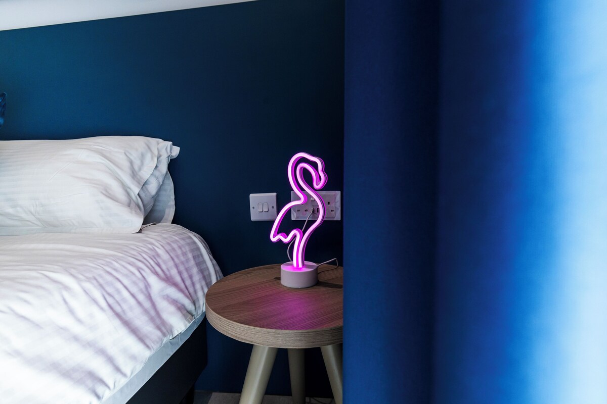 A bedroom with a bed, a round wooden table, and a neon flamingo sign.