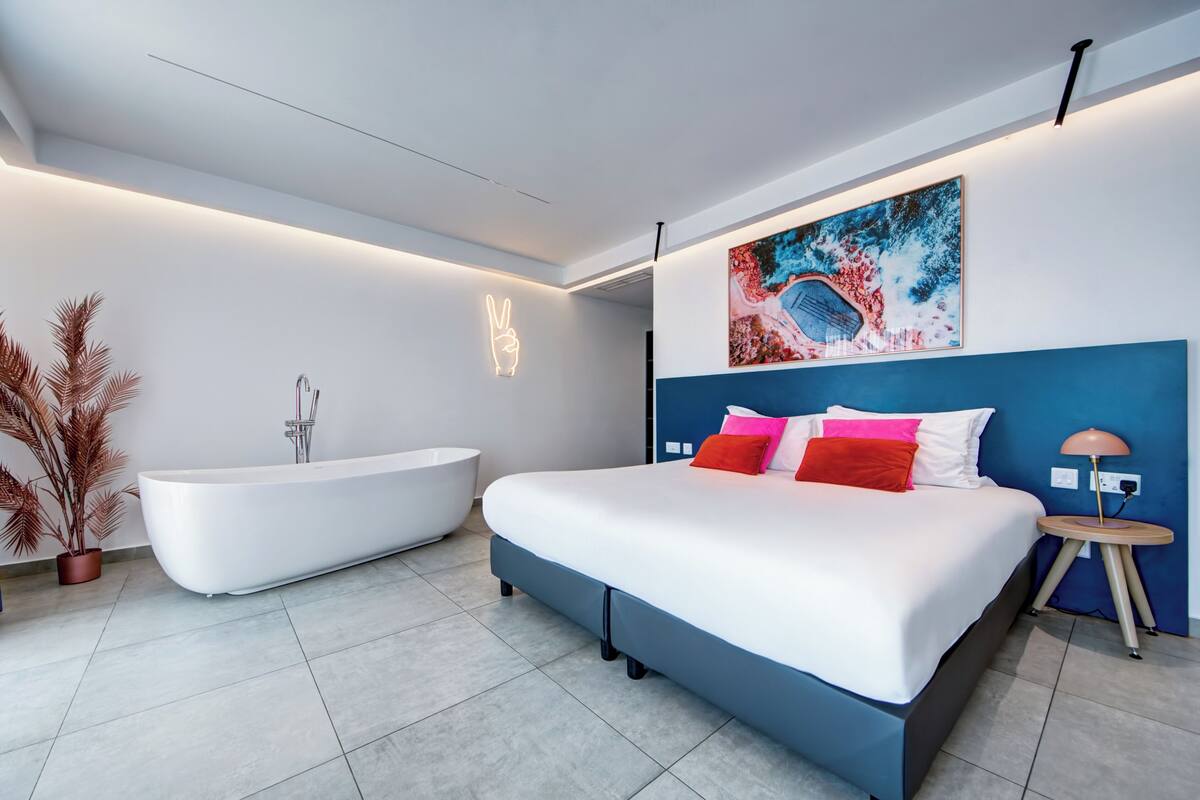 A modern bedroom with a large bed, a white bathtub, and a wall-mounted light.