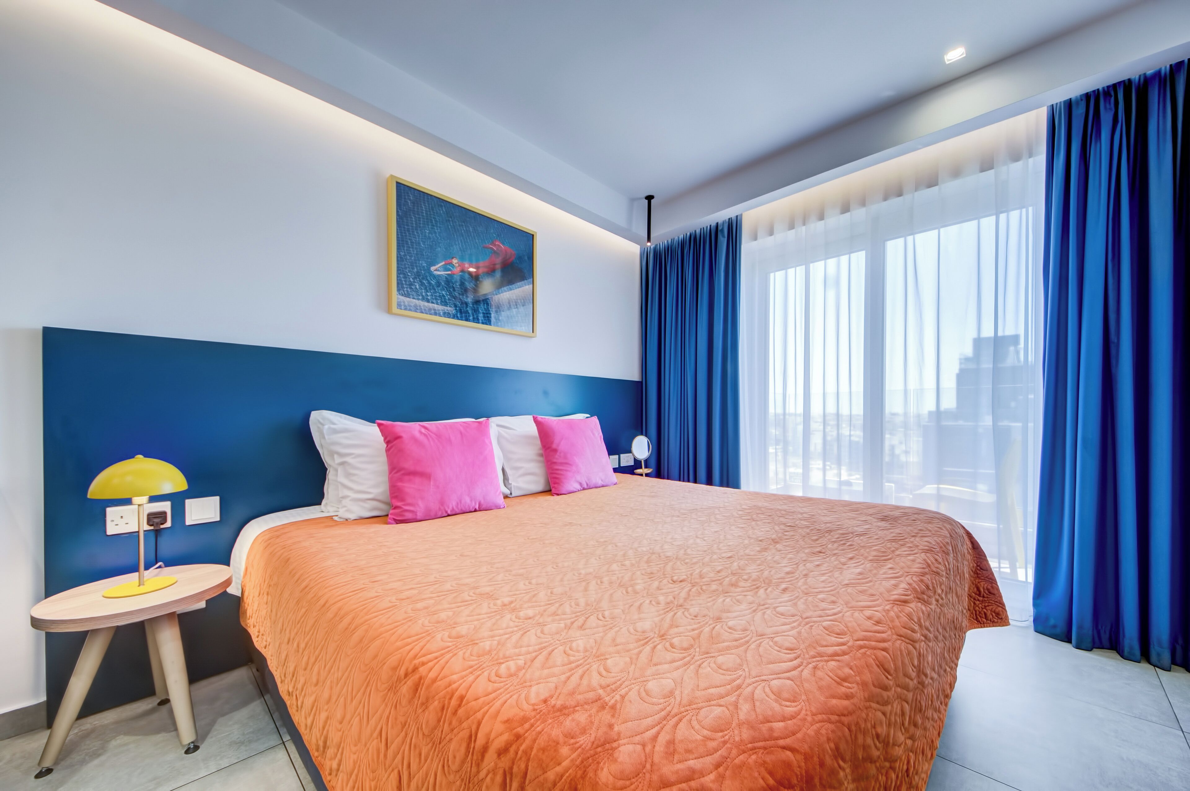 A hotel room with a large bed, a nightstand, a lamp, a painting of a swimmer, and a view of the city through large windows.