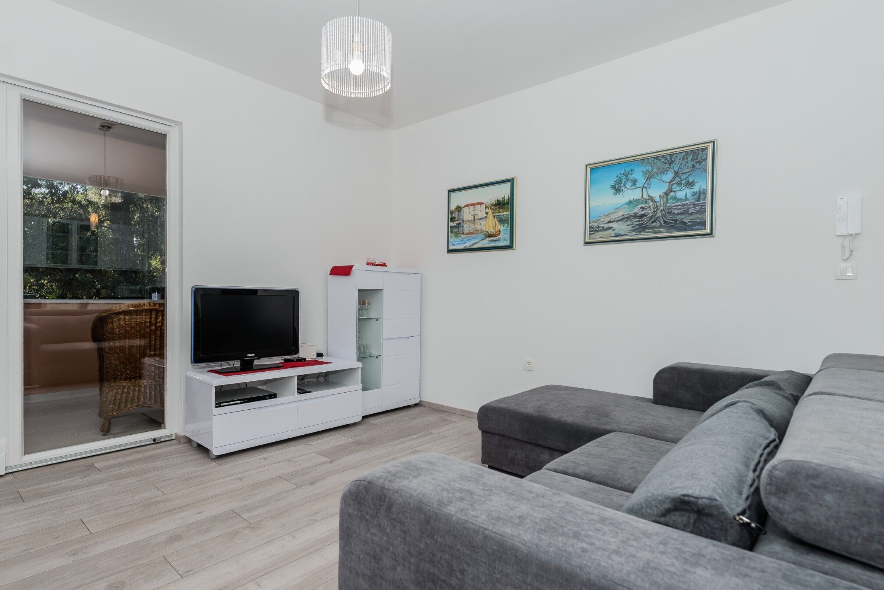 A modern living room with a grey sofa, a flat-screen TV on a white entertainment unit, and two framed landscape paintings on the wall.