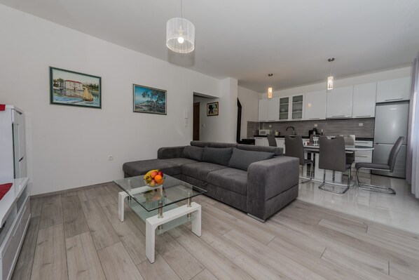 Una sala de estar moderna con un sofá gris, una mesa de centro de vidrio con fruta, y una cocina al fondo.