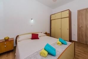 A neatly made bed with a red pillow, a wooden nightstand with a lamp, and a wardrobe with a wooden door. - Spacious Apartment w Patio 40m From the Beach (Kastela)