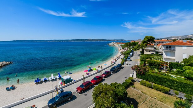 Una carretera costera con coches aparcados, una playa con sombrillas y edificios a lo largo de la orilla.