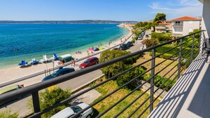 Vista costera desde un balcón con vista a una playa, coches estacionados a lo largo de la carretera, cielo despejado y edificios al fondo.