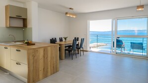 Una cocina moderna con una isla de madera, un área de comedor con sillas y un amplio ventanal corredera que da a un balcón con vistas al mar.