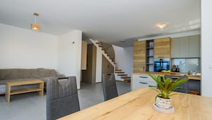 Una sala moderna con un sofá, una mesa de centro de madera y una mesa de comedor con una planta en maceta. Una escalera conduce a una zona de cocina con armarios de madera y un horno empotrado.