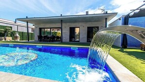 En moderne udendørs swimmingpool med et vandfald, omgivet af en velplejet græsplæne og en moderne bygning med store vinduer.