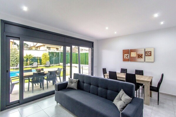 A modern living room with a dark grey sofa, a wooden dining table with chairs, and a large sliding glass door leading to an outdoor patio with a pool and garden furniture.