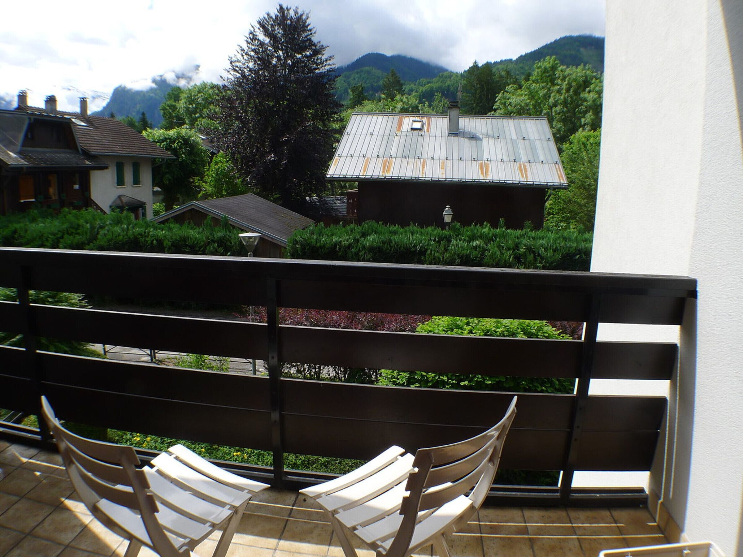 A balcony with two white lounge chairs, a view of a mountain, and a building with a metal roof.