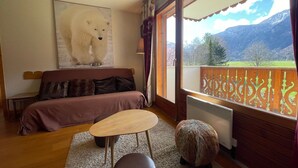 A cozy room with a bed, a small table, and a sofa. There's a large window with a view of mountains and a balcony with a decorative railing.