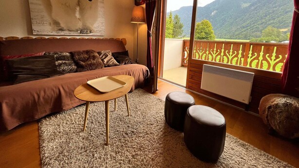 A cozy room with a sofa, a small table, and two ottomans. There's a large window with a view of mountains.