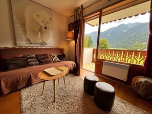 Een gezellige kamer met een bank, een klein tafeltje en twee poufs. Er is een groot raam met uitzicht op bergen.