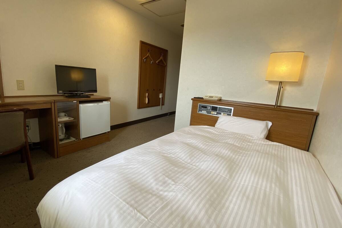 A hotel room with a bed, a desk with a television, a refrigerator, a chair, a wardrobe, and a bedside lamp.