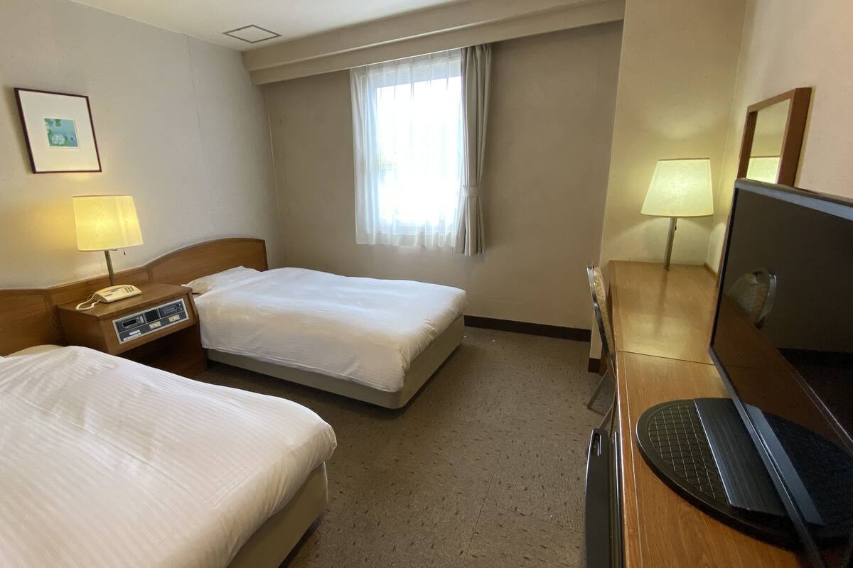A hotel room with two beds, a desk with a computer, a TV, a window with curtains, and a bedside lamp.