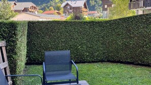 A well-maintained garden with a metal chair and a wooden bench, surrounded by neatly trimmed hedges and a view of mountain houses.