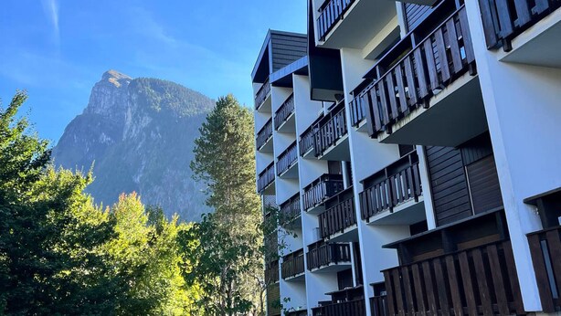 A multi-story building with balconies, surrounded by trees and a mountain in the background.