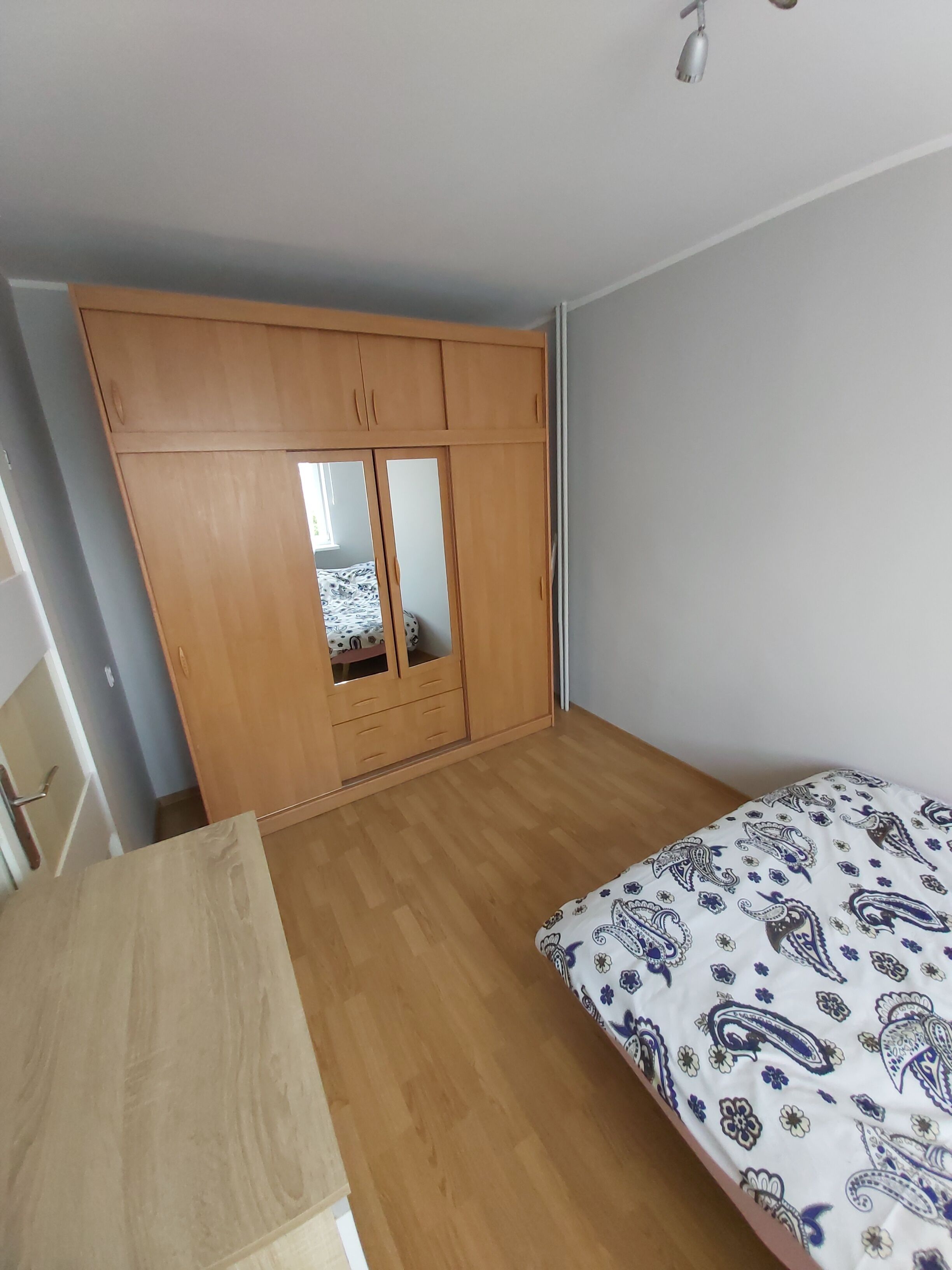 A bedroom with a wooden wardrobe, a bed with a patterned duvet, and a wooden desk.